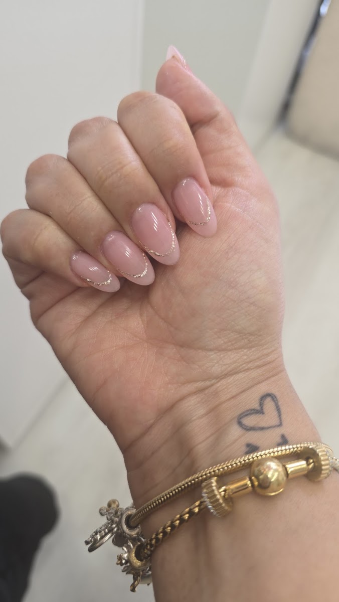 Mano con uñas ovales rosadas con borde blanco y detalles dorados, junto a un tatuaje de corazón en la muñeca